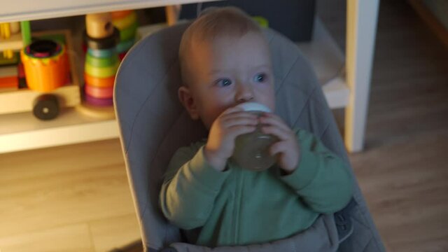 Little cute baby drinks juice through a pacifier from baby bottle in front of TV, caucasian toddler watching tv in dark living room. High quality 4k footage