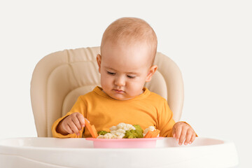 A newborn child aged one year tries boiled vegetables for the first time. A baby carefully takes a boiled carrot while sitting on a feeding chair on a white background