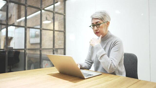 Mature asian business woman wears glasses using laptop computer sit at workplace desk. Happy senior older employee 50s businesswoman