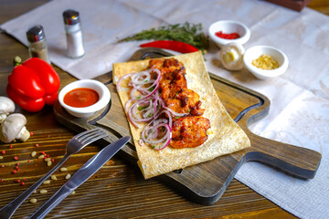 On a dark tabletop, a light linen napkin, on a cutting board in pita bread there is a ready-made shish kebab with rings of red onion, cutlery, a couple of salt and pepper, a pod of hot pepper