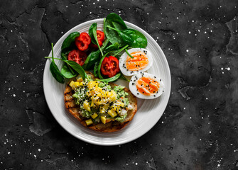 Fototapeta na wymiar Delicious healthy breakfast, snack - sandwich with avocado, shrimp, mango salsa, boiled egg and spinach tomatoes salad on a dark background, top view