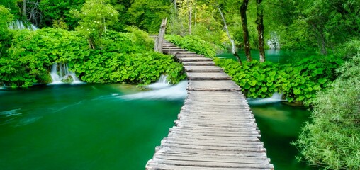 Long exposure scene with a rustic wooden boardwalk in a beautiful green forest, crossing ponds with small waterfalls and lush green summer foliage, in Plitvice Lakes National Park, Croatia.