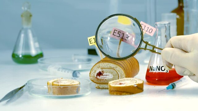 The inspector examines the sweet roll from the store in the laboratory with a magnifying glass. Sweet roll on the table is decorated with plates with the names of additives E. Food safety laboratory, 