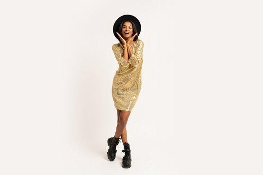 Studio photo of beautiful black woman in golden party dress posin on white background. Wearing stylish hat and earrings.  Full lenght.