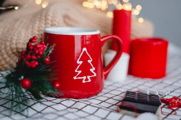 Obraz na płótnie Canvas Red mug with coffee in Christmas decorations on a white checkered blanket. New year, garlands, gifts, candles - an atmosphere of warmth, comfort and magic.