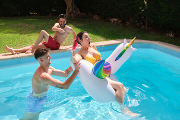 Young man throwing woman from a rainbow unicorn float into the swimming pool.