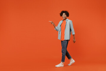 Full body young student black man 50s wearing blue shirt t-shirt walk go point index finger aside on copy space area isolated on plain orange color background studio portrait People lifestyle concept