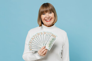 Elderly smiling cheerful fun happy woman 50s wear white knitted sweater holding fan of cash money in dollar banknotes isolated on plain blue color background studio portrait. People lifestyle concept.