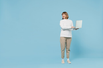 Full body elderly smiling woman 50s wearing white knitted sweater hold use work on laptop pc computer look aside on copy space area isolated on plain blue color background. People lifestyle concept.