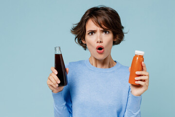 Young shocked woman 20s wear casual sweater hold soda cola water vegetable smoothie choose between junk food and healthy isolated on plain pastel light blue background. People lifestyle food concept.