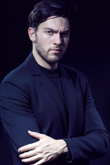 Serious Handsome Caucasian Brunet Businessman Wearing Black Suit With Folded Hands Posing With Straight Look Against Black Studio Background.