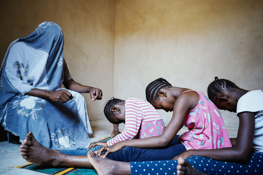Totally covered person holding a blade speaking to a group of intimidated black African girls sitting in a row on the floor with their heads bowed prior to the female genital mutilation intervention