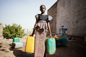 Pretty black African girl with two large water canisters leaving the public water place on a hot...