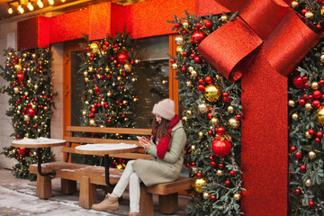 woman sitting at cafe terrace decorated for christmas, using mobile phone for texting or social media