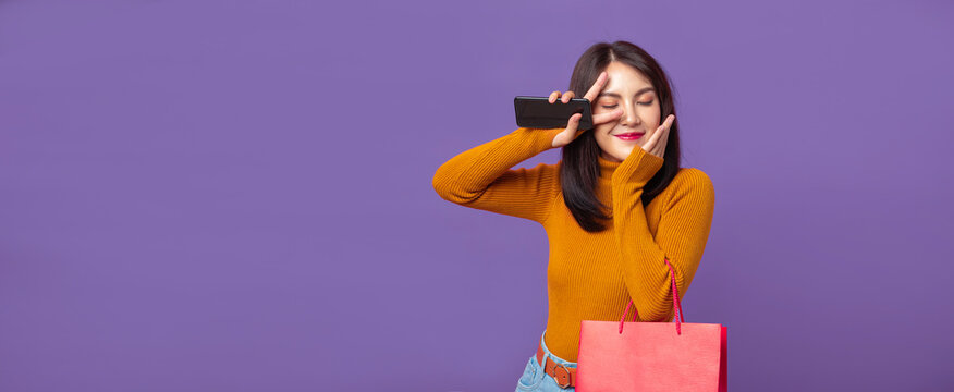 Cheerful Young Beautiful Asian Female In Fashion Yellow Long-sleeve Shirt With Bag Shopping Online With Mobile Phone Isolated On Purple Banner Background With Copy Space.