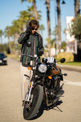 Adolescente hombre joven motero con camisa de cuadros elegante preparándose para viaje. Fondo de palmeras veraniegas. Chico elegante con gafas de sol y moto. Chico poniéndose casco de seguridad.