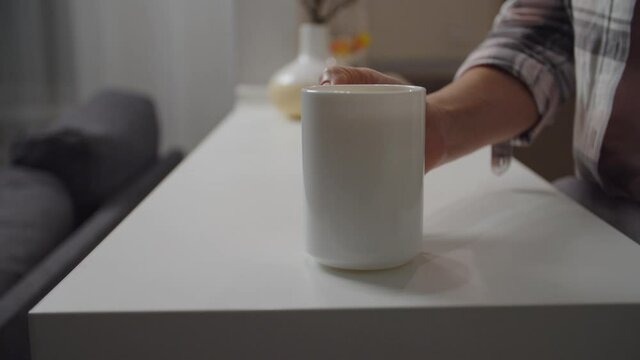 Unrecognizable person putting white mug with drink on kitchen counter indoors. Focus on foreground. Blurred background of domestic room. Advertisement concept of hot beverage, coffee, tea, cacao