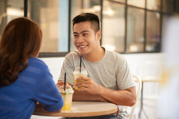 Smiling young Asian man in tshirt sitting at table and drinking lemonade while dating with girl in cafe