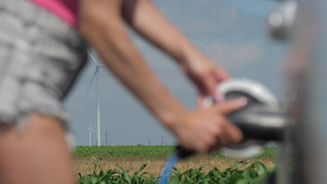Wind machines generate electricity in field. Young woman disconnects charger off electric car at charging station on summer closeup