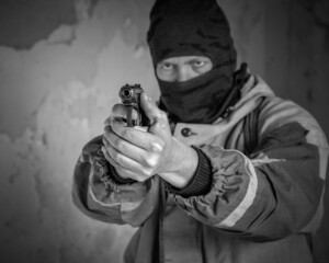 An armed robber in a mask and a pistol in his hands prepares to fire. Armed robbery, black and white photo, selective focus