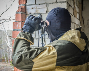 A masked soldier armed with a Kalashnikov assault rifle with a silencer looks through binoculars....