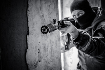 A masked soldier armed with a silenced assault rifle is about to fire. Armed attack, black and white photo, selective focus. The special police are conducting a counter-terrorist operation.  - obrazy, fototapety, plakaty
