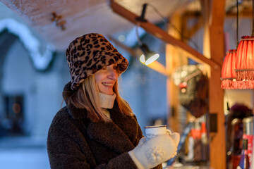 Woman in fur coat, panama hat at Tallinn Christmas market drinking hot mulled wine from reusable cup