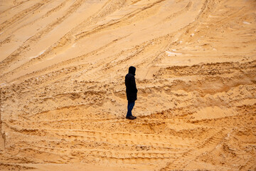 Fototapeta na wymiar tall slim man from his back in black elongated jacket with hood on his head walks in the sand in quarry between the sandy holly and dunes, with trails from tractors and cars.
