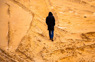Fototapeta premium tall slim man from his back in black elongated jacket with hood on his head walks in the sand in quarry between the sandy holly and dunes, with trails from tractors and cars.
