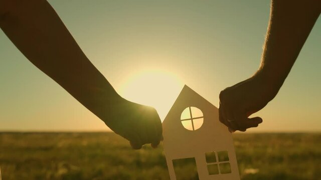 The concept of building a house for the family. The family is holding a paper house in their hands, at sunset, the sun is shining through the window. Dream to buy a house. Symbol of home, happiness
