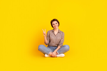 Full size photo of young attractive woman good mood sit floor show fingers v-symbol isolated over yellow color background