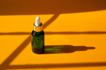 Green glass dropper bottle with metallic lid. Natural sunshine and shadows from tropical plants. Orange background with daylight. Skincare products , natural cosmetic. Beauty concept for face body