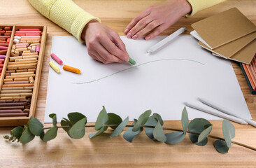 Woman drawing eucalyptus branch with soft pastels at wooden table, closeup