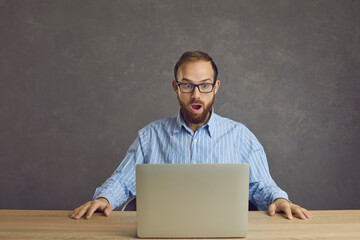 Man sitting at a table with a laptop on a gray background is in great shock from what he saw on the screen. Adult bearded man with glasses looks at the shocking content or gets a good job offer.