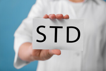 Doctor holding sheet of paper with abbreviation STD on light blue background, closeup