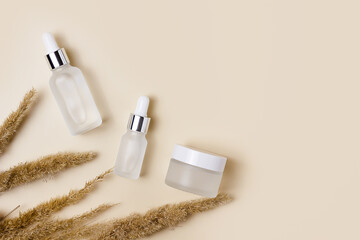 White cosmetic jars on a beige background with dry spikelets of field grass. Winter and autumn care products. Cream, serum, fluid. Minimalism and sustainability. 