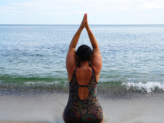 woman in a swimsuit performs yoga while sitting on the shore at the water's edge