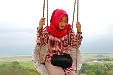 Hijab woman enjoy the views on the swing from the top of the hill in Eling Bening in Ambarawa,Central Java,Indonesia with mountain and cloudy sky background.