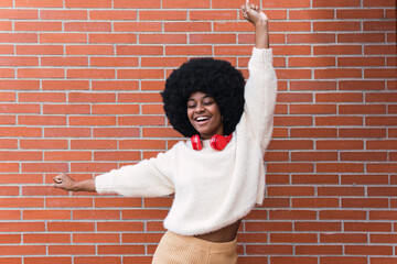 Cheerful african american woman with red headphones on neck, white perfect smile, long afro hair and white jumper dancing and having fun in brick wall. Copy space