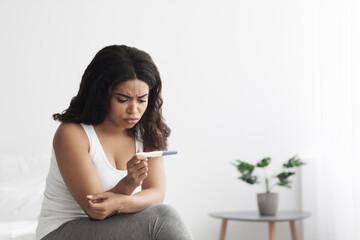 Upset african american woman checking her recent pregnancy test, sitting on bed at home, free space