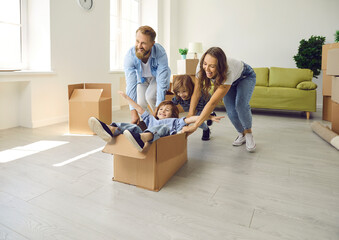 Fototapeta na wymiar Husband and wife with children who have just moved into their own house are having fun and rejoicing. Parents ride their little sons in cardboard box. Concept of buying your own home and moving.