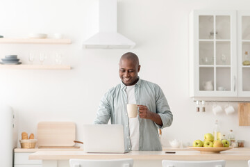 Modern worker in self-isolation. Happy mature african american man with cup, drinking coffee and working on laptop