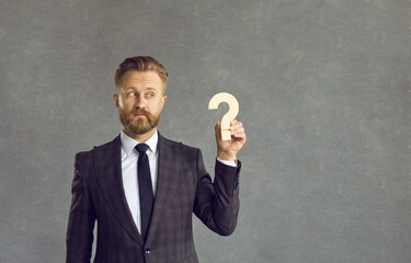 Confused puzzled businessman holding wooden question mark standing on gray on background. Pensive...