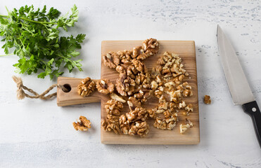 Wooden board of chopped walnuts and greens with a knife on a light gray textured background, home cooking