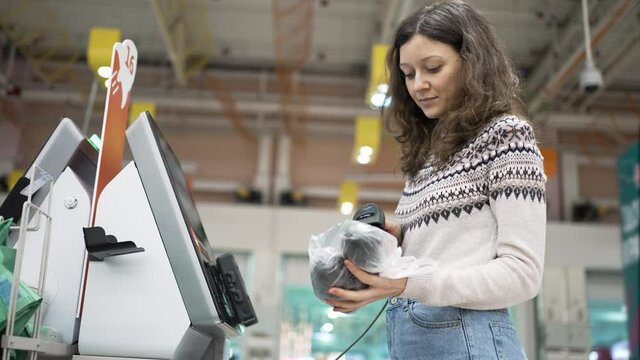 A young woman at a self-service checkout in a grocery supermarket makes purchases, punches barcodes on the product and pays