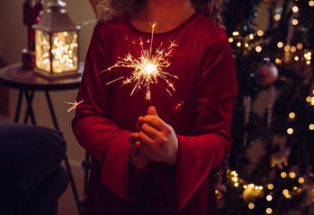 Cute girl child holding sparkler candle in hands in home living room, decorated Christmas tree on background, dark room, lit by sparkler candle. Happy New Year greetings.