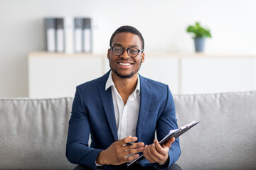 Portrait of happy black male psychologist looking at camera and taking notes during therapy session at clinic - obrazy, fototapety, plakaty