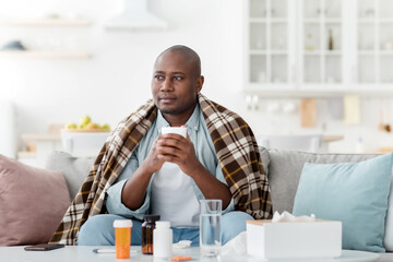Thoughtful african american man feeling sick, holding mug with hot drink, sitting on sofa near table full of pills
