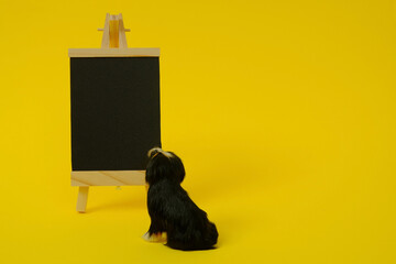 A black board with a wooden stand and a figure of a black dog looking at the board on a yellow...