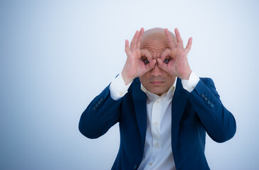 A bald man in a business suit depicts binoculars with a hand gesture. Stylish businessman looks through imaginary binoculars. Concept: curiosity and interest